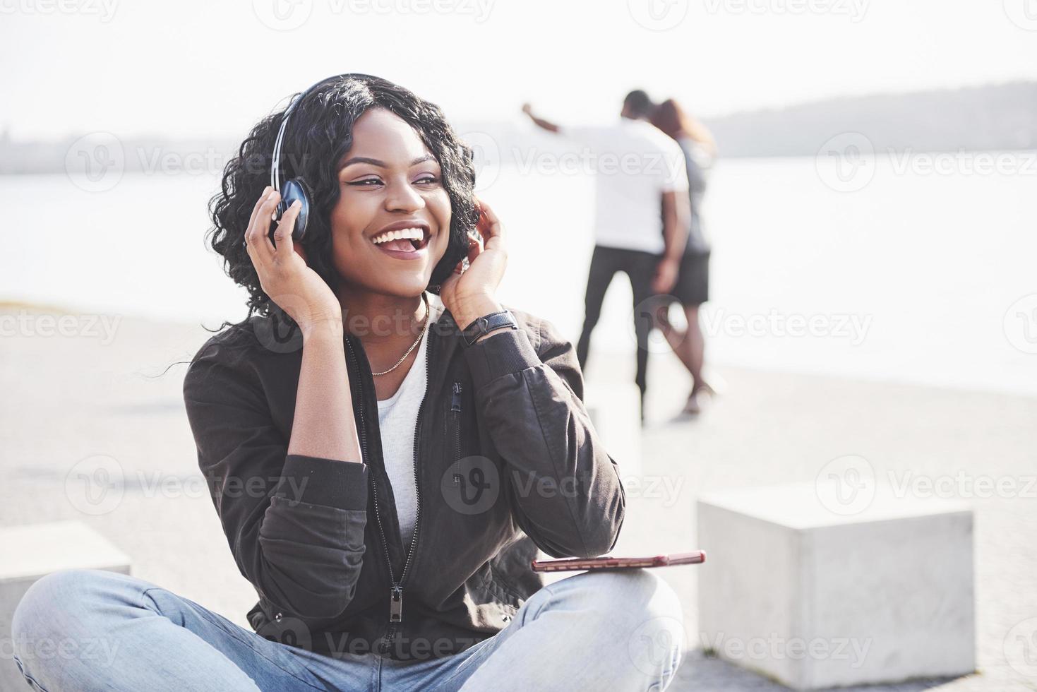 porträtt av en vacker ung söt afroamerikansk flicka som sitter på stranden eller sjön och lyssnar på musik i sina hörlurar foto