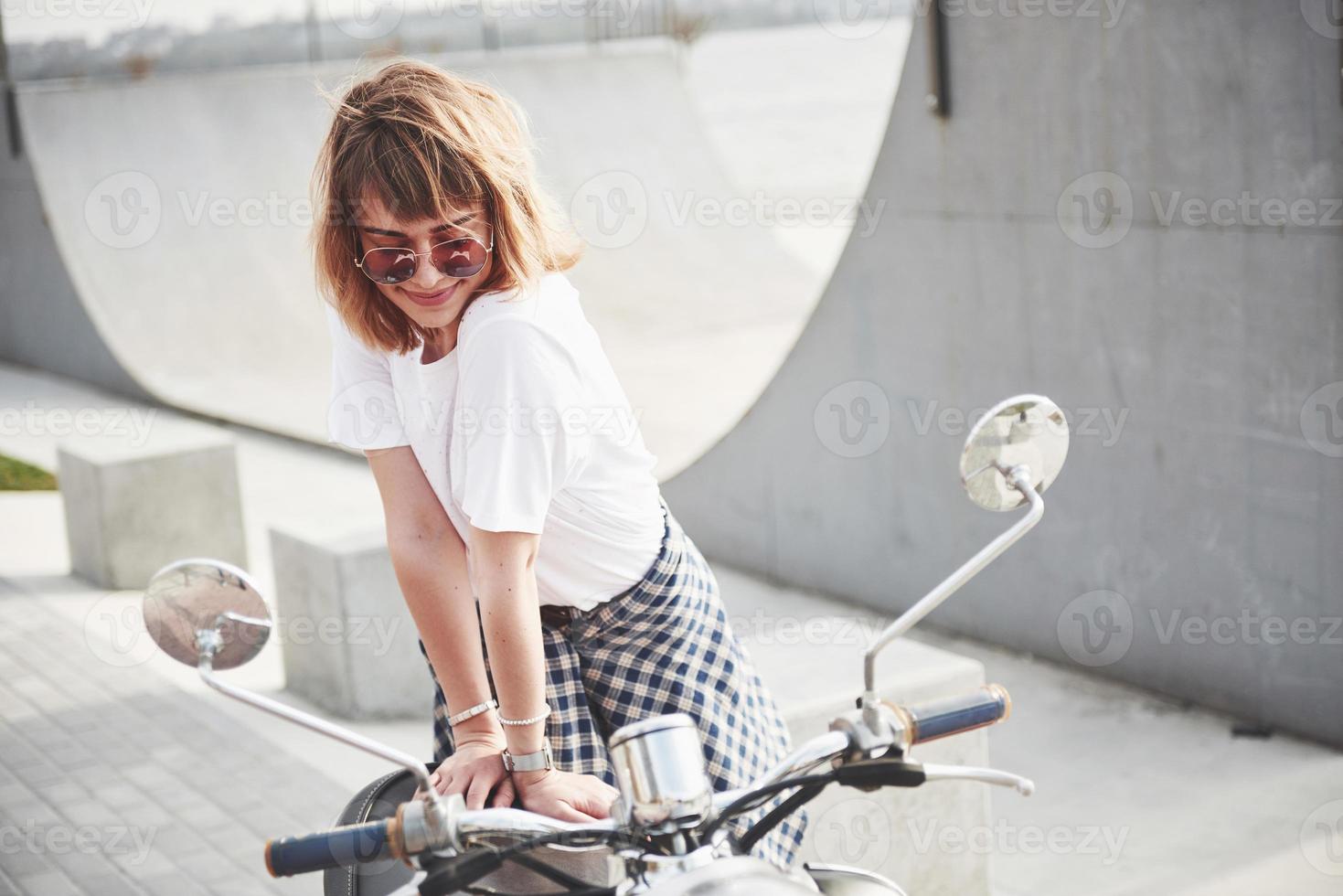 porträtt av en vacker flicka hipster sitter på en svart retro scooter, leende poserar och njut av det varma vårsolen. foto