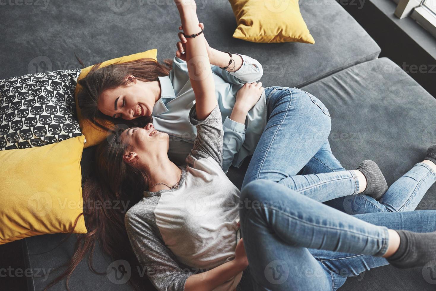 två söta leende tvillingar systrar håller smartphone och gör selfie. flickor ligger i soffan och poserar och glädjer sig foto