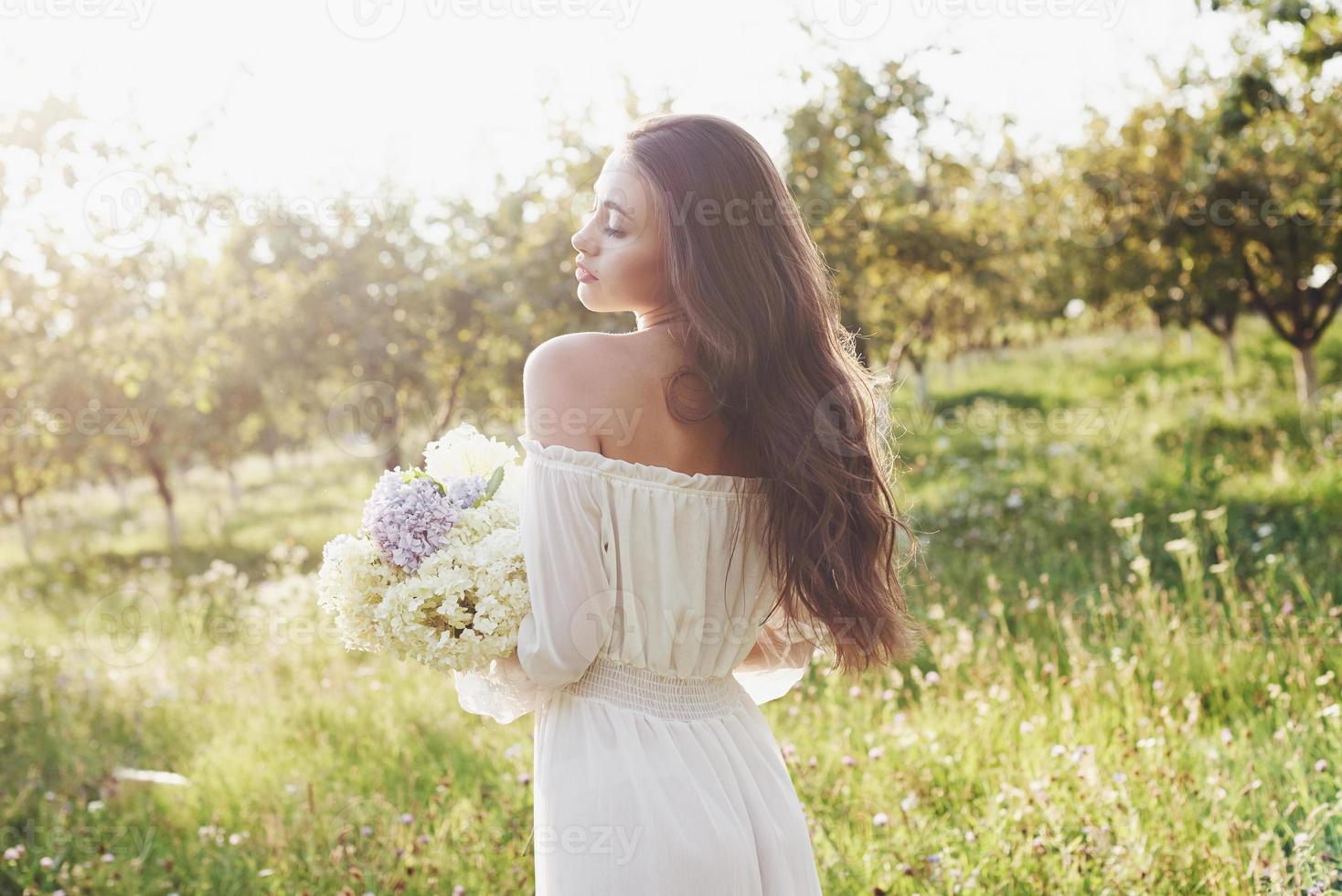 en vacker ung flicka i en vit ljus klänning och en bukett sommarblommor lägger en fin dag i trädgården foto