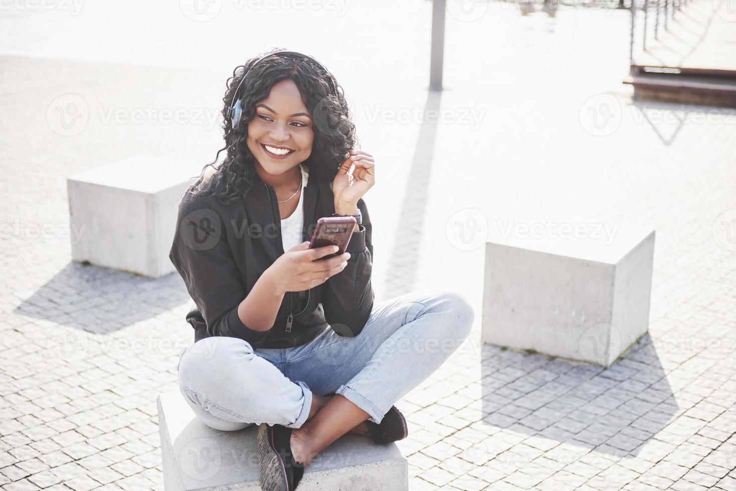 porträtt av en vacker ung söt afroamerikansk flicka som sitter på stranden eller sjön och lyssnar på musik i sina hörlurar foto