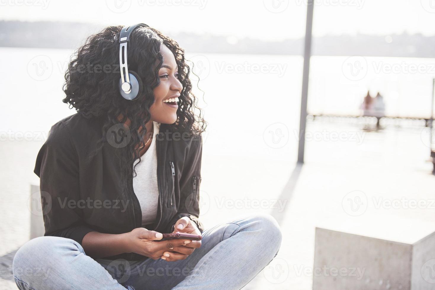 porträtt av en vacker ung söt afroamerikansk flicka som sitter på stranden eller sjön och lyssnar på musik i sina hörlurar foto