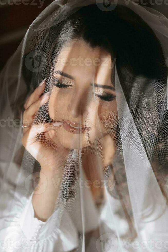 de brud är framställning för de bröllop. en drömmande flicka i en klä på sig klänning med lång mörk hår nära de fönster under en slöja foto