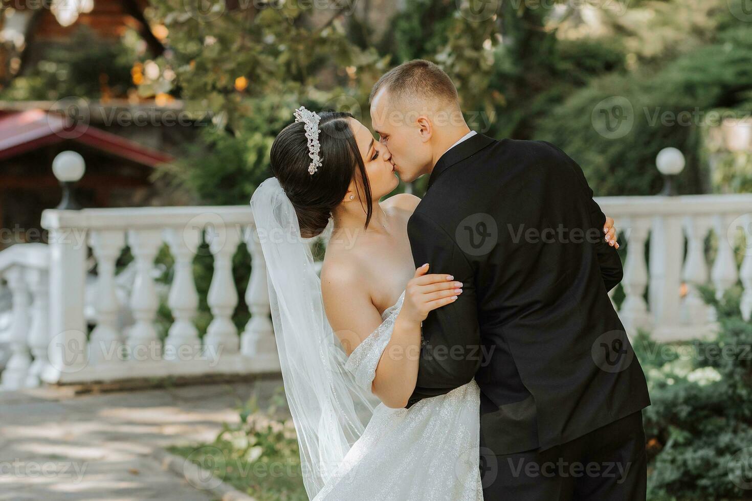 porträtt av en ung skön bröllop par i de trädgård, de brud i ett utanför axeln bröllop klänning, de brudgum i en svart klassisk kostym, ömt kissing på de bakgrund av natur foto