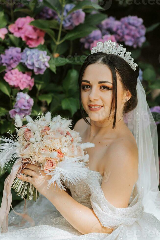 ung skön brud i bröllop klänning med öppen axlar och krona på henne huvud Sammanträde nära hortensia blommor, mode Foto tagen under hård solljus