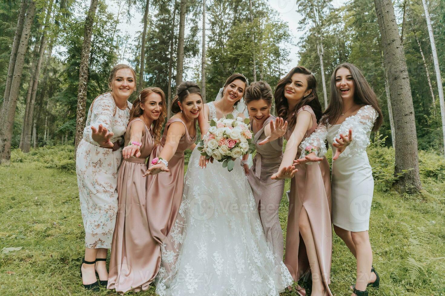 en grupp av skön flickor med en brud i identisk klänningar är leende, fira och har roligt tillsammans mot de bakgrund av natur och lång träd. flickor fest foto