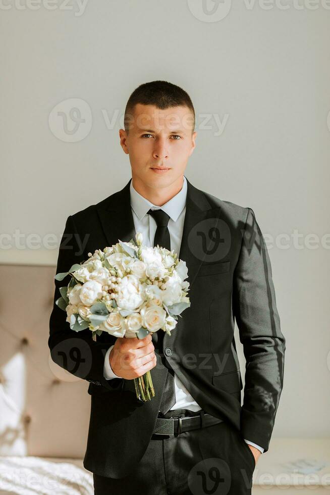 eleganta brudgum med en bukett av blommor i en hotell rum. brudgummens morgon. de brudgum är få redo i de morgon- innan de bröllop ceremoni foto