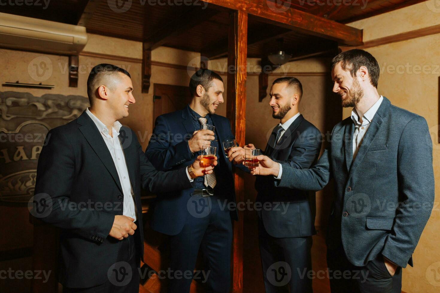 de brudgum och hans vänner i eleganta kostymer dryck whisky i de hotell rum, de morgon- innan de bröllop förberedelser. grupp av män talande och dricka whisky foto