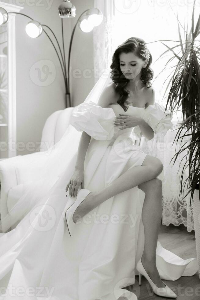 skön modell flicka med lång lockigt hår i en modern bröllop klänning. vertikal porträtt. en skön brud i en vit bröllop klänning bär högklackade skor. svart och vit Foto
