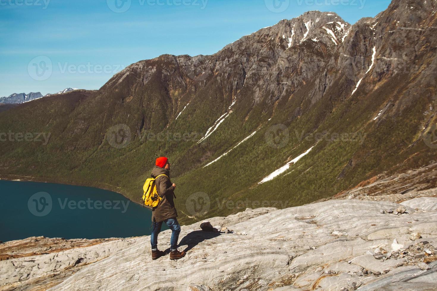 man med en ryggsäck som står på en sten på bakgrund av berg och sjö foto