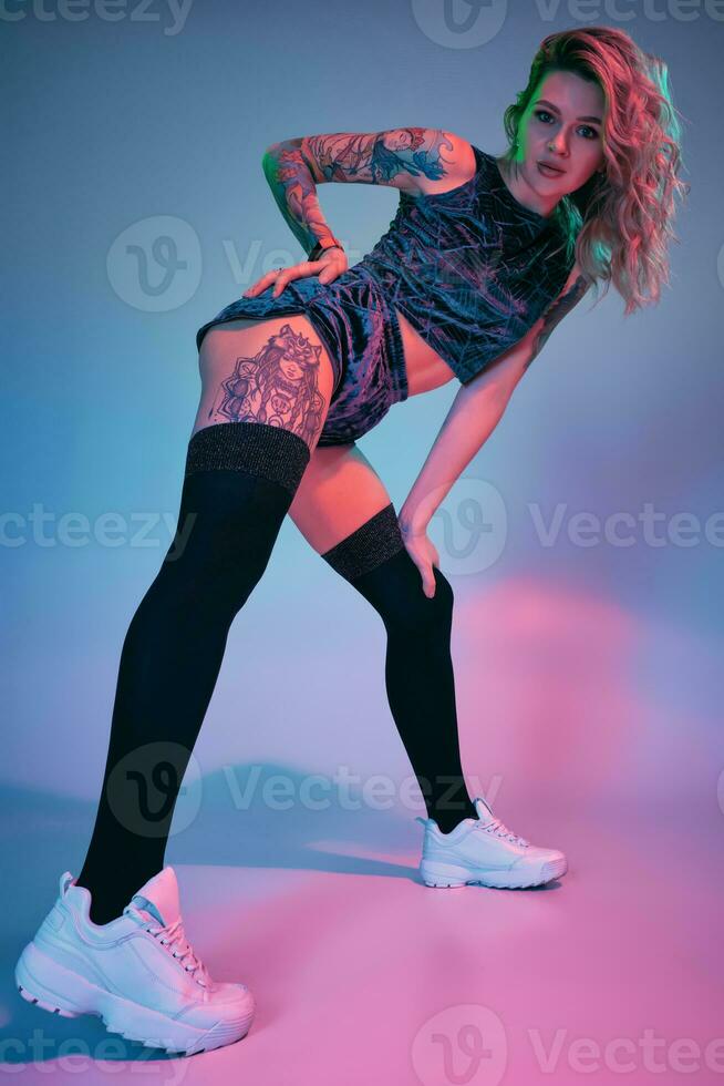 ung blond kvinna i velour blå sport ha på sig och svart strumpor förföriskt Framställ på kamera i studio. rosa och blå bakgrund foto