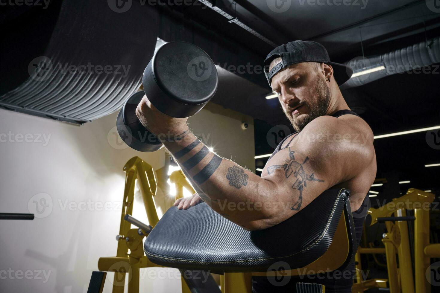 tatuerade kille med örhänge, i svart väst och keps. han är lyft en hantel, Träning hans biceps, Sammanträde på predikant ringla bänk på mörk Gym. stänga upp foto