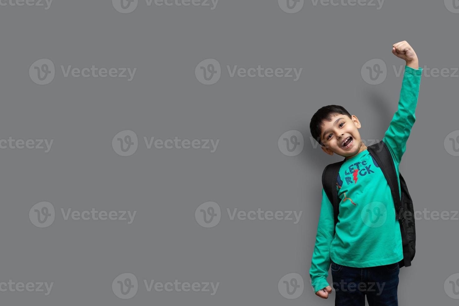 söt 7-årig glad pojke med skolryggsäck som håller böcker i vanlig bakgrund för utbildningskoncept foto