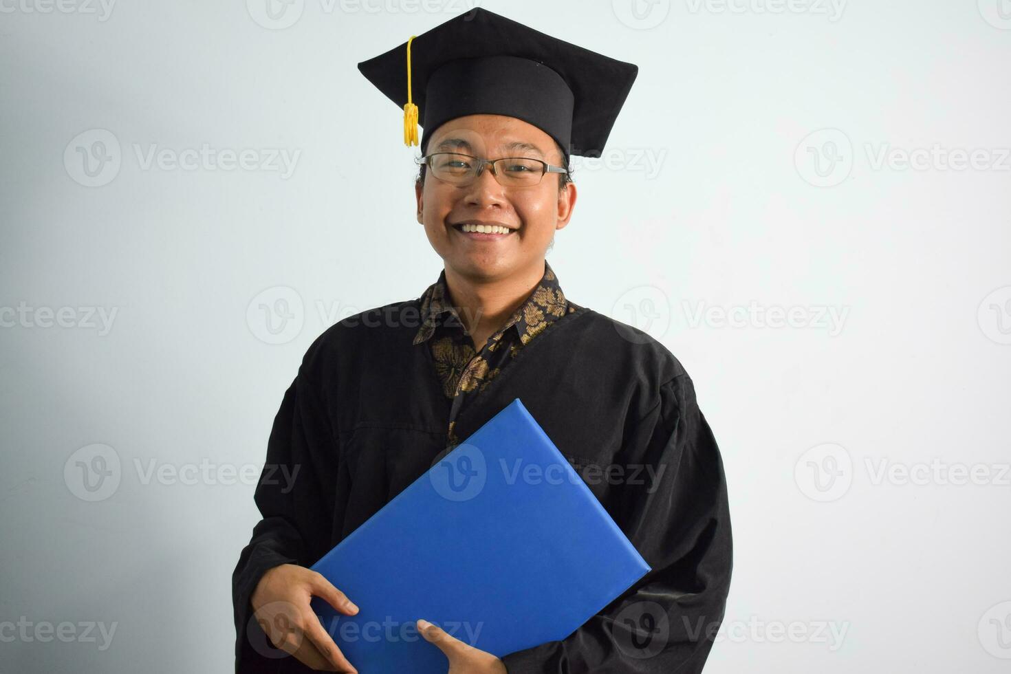 uttrycksfull av vuxen indonesien manlig ha på sig gradering morgonrock, hatt och glasögon, asiatisk manlig gradering föra tom blå certifikat isolerat på vit bakgrund, uttryck av porträtt gradering foto