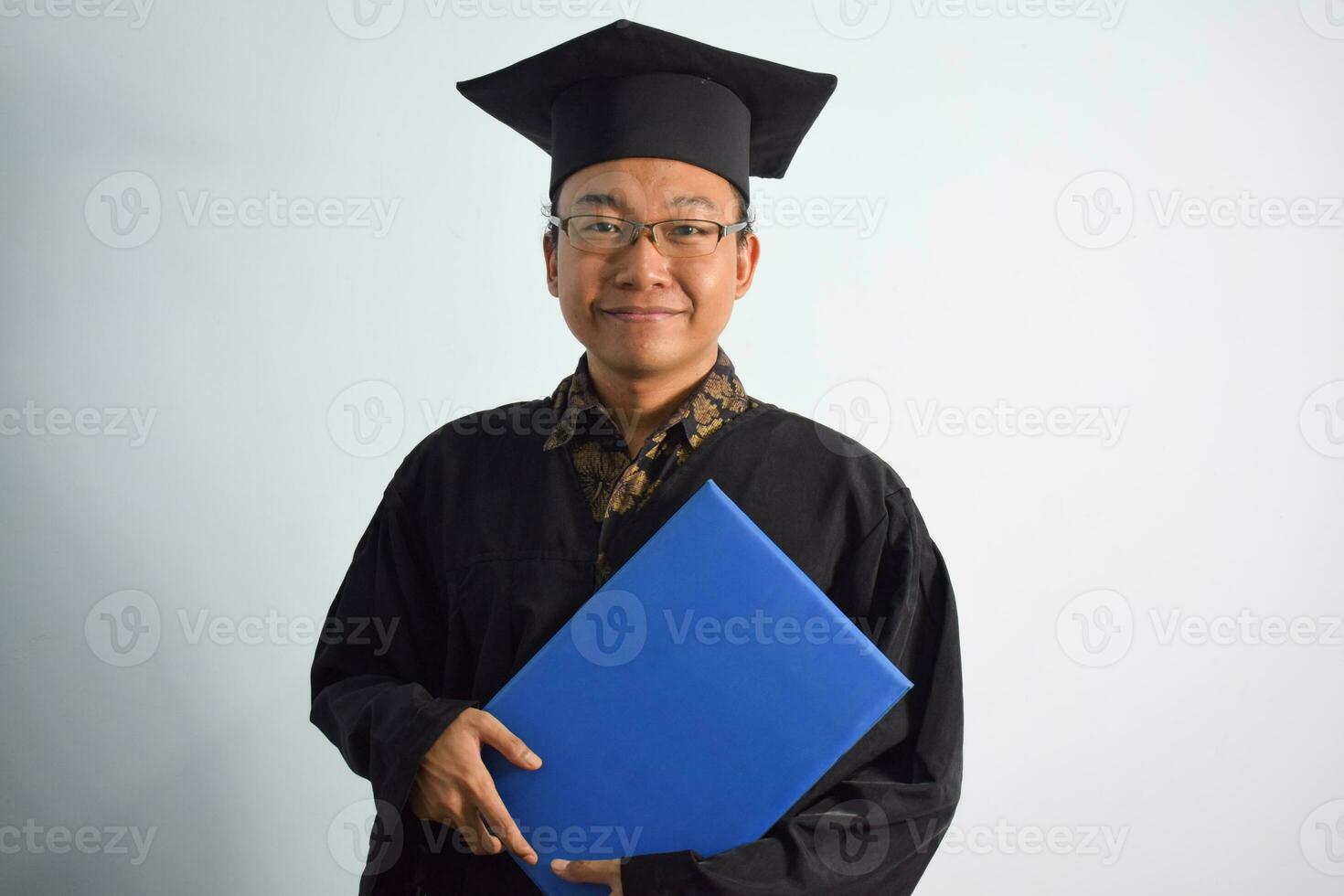 uttrycksfull av vuxen indonesien manlig ha på sig gradering morgonrock, hatt och glasögon, asiatisk manlig gradering föra tom blå certifikat isolerat på vit bakgrund, uttryck av porträtt gradering foto