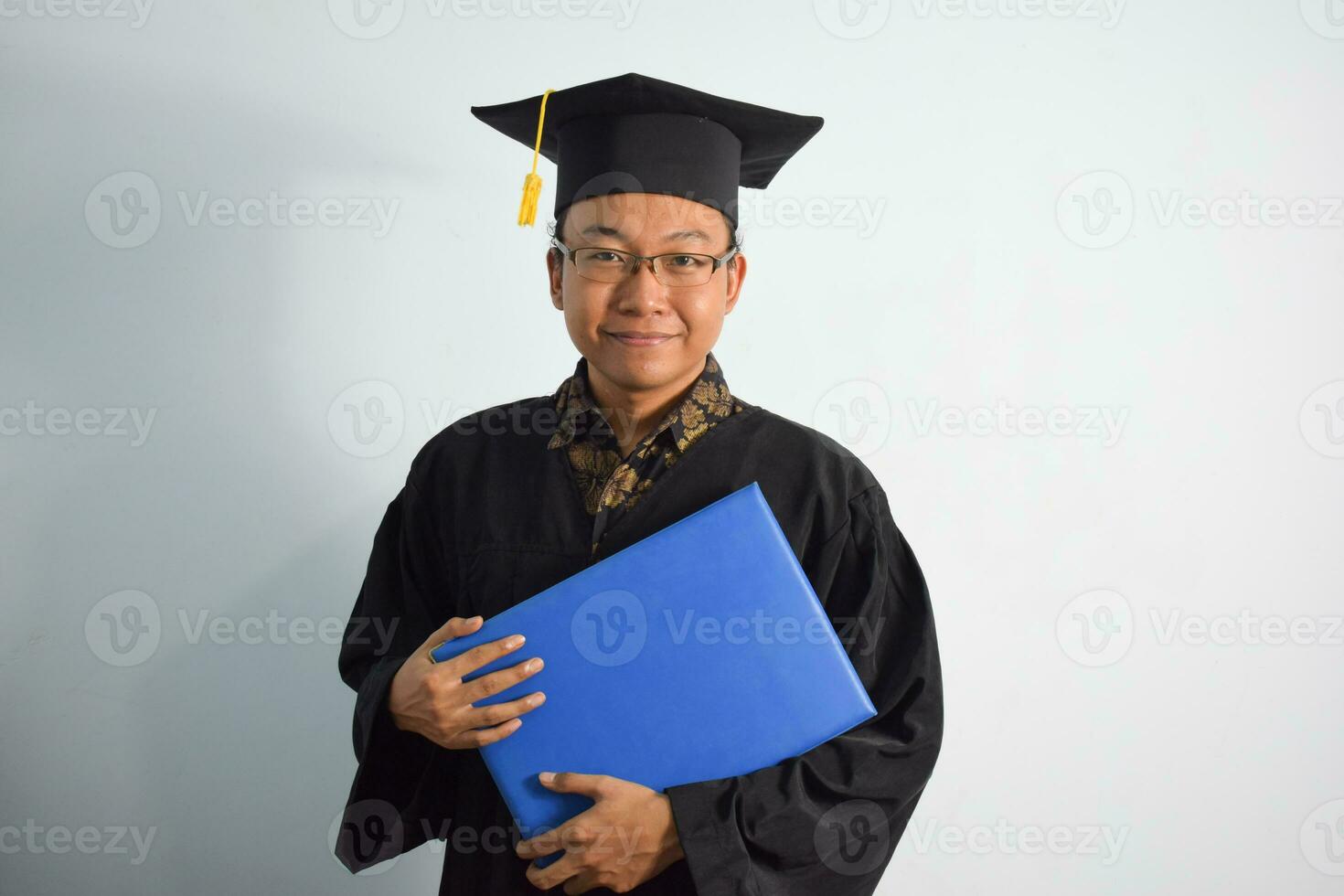 uttrycksfull av vuxen indonesien manlig ha på sig gradering morgonrock, hatt och glasögon, asiatisk manlig gradering föra tom blå certifikat isolerat på vit bakgrund, uttryck av porträtt gradering foto