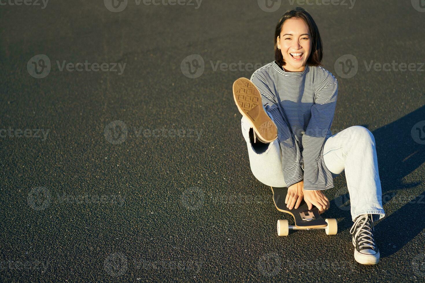 porträtt av skön asiatisk flicka skridskoåkning, Sammanträde på henne skateboard och leende. söt tonåring med longboard, skridskoåkning på väg foto