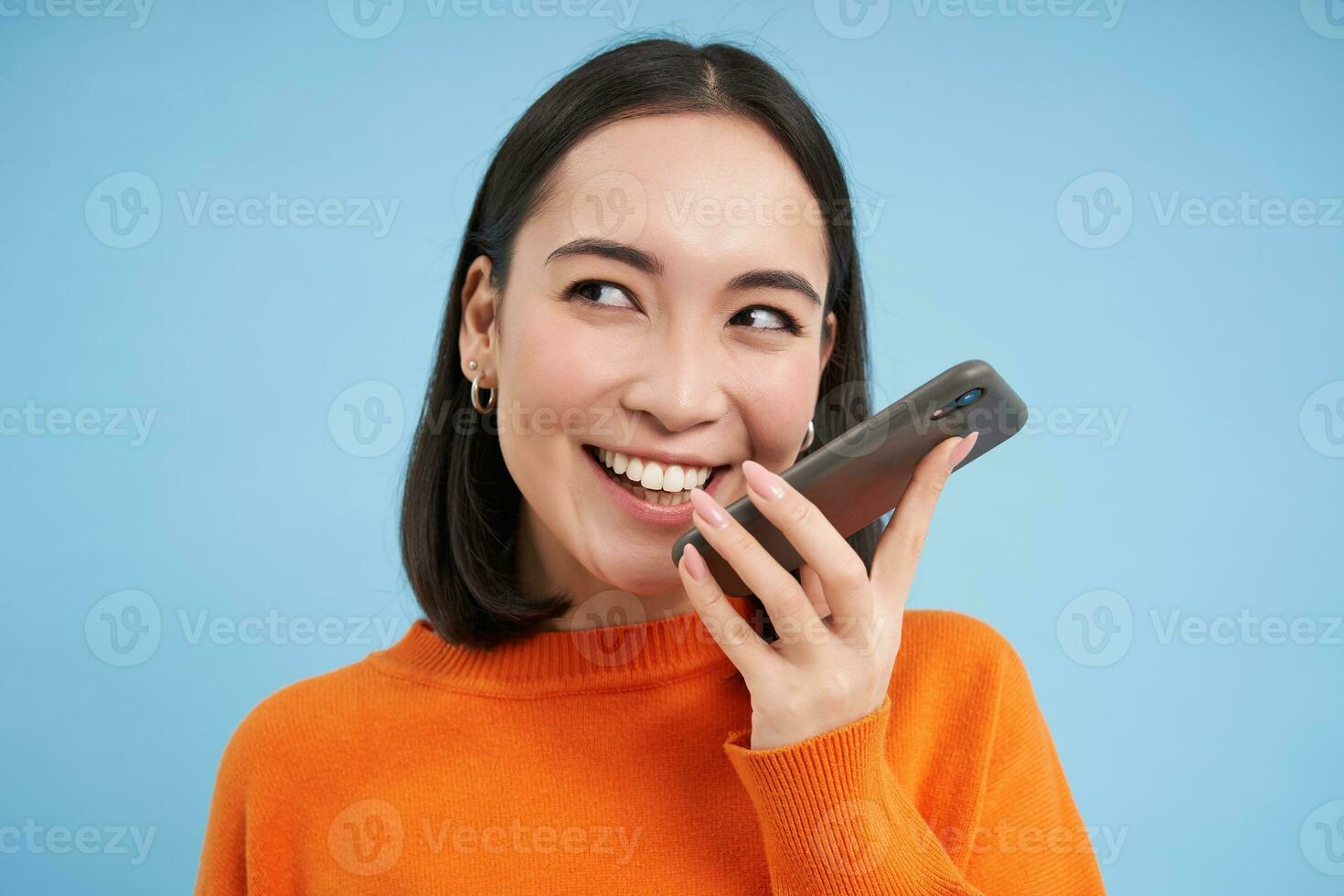 porträtt av asiatisk kvinna uppgifter röst meddelande, samtal på högtalartelefon, innehar mobil telefon nära mun medan tala, översätter henne Tal på app, blå bakgrund foto