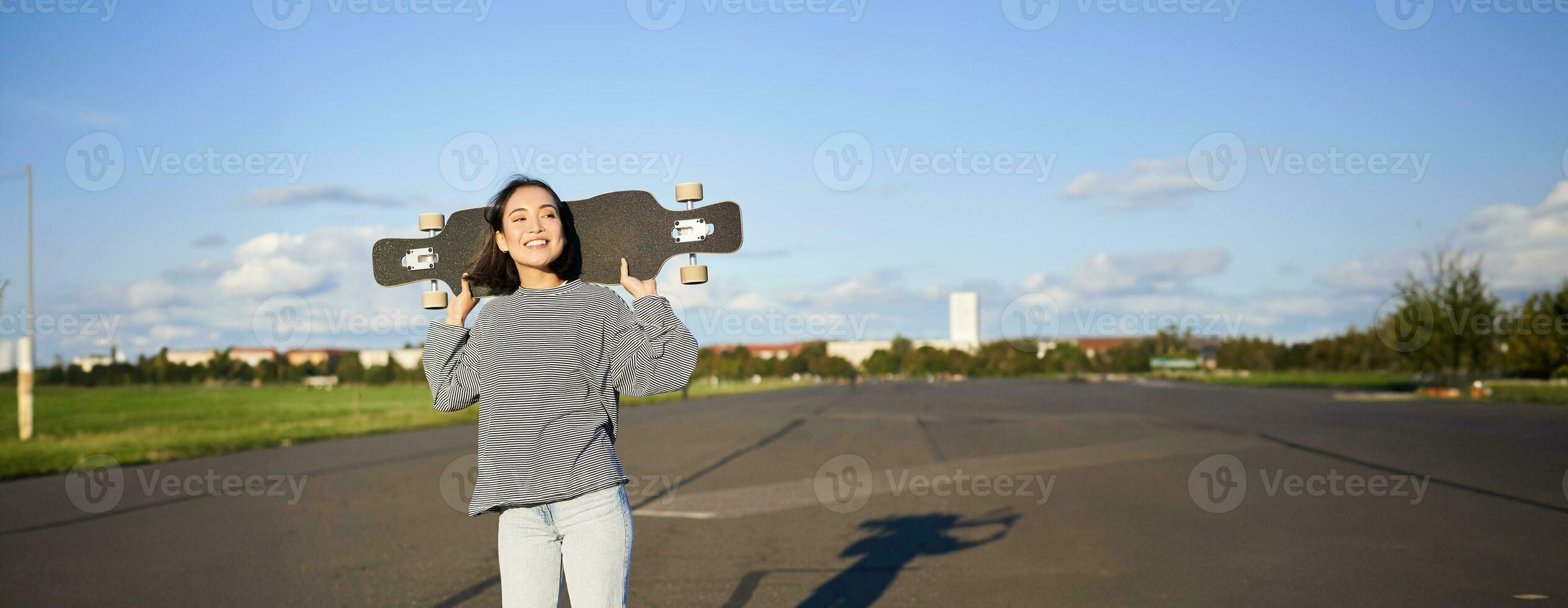 livsstil och människor. ung asiatisk flicka Framställ med longboard, skridskoåkning på henne kryssare. leende kvinna innehav skateboard på axlar foto