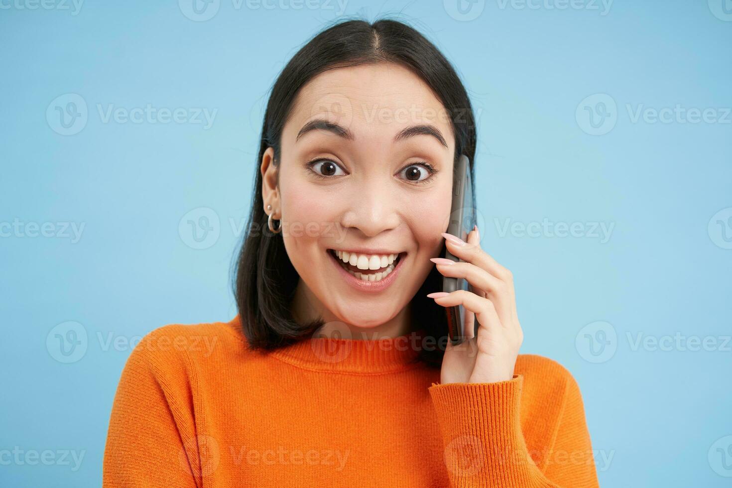 glad asiatisk flicka samtal på mobil telefon, talar med någon på telefon, står över blå bakgrund foto