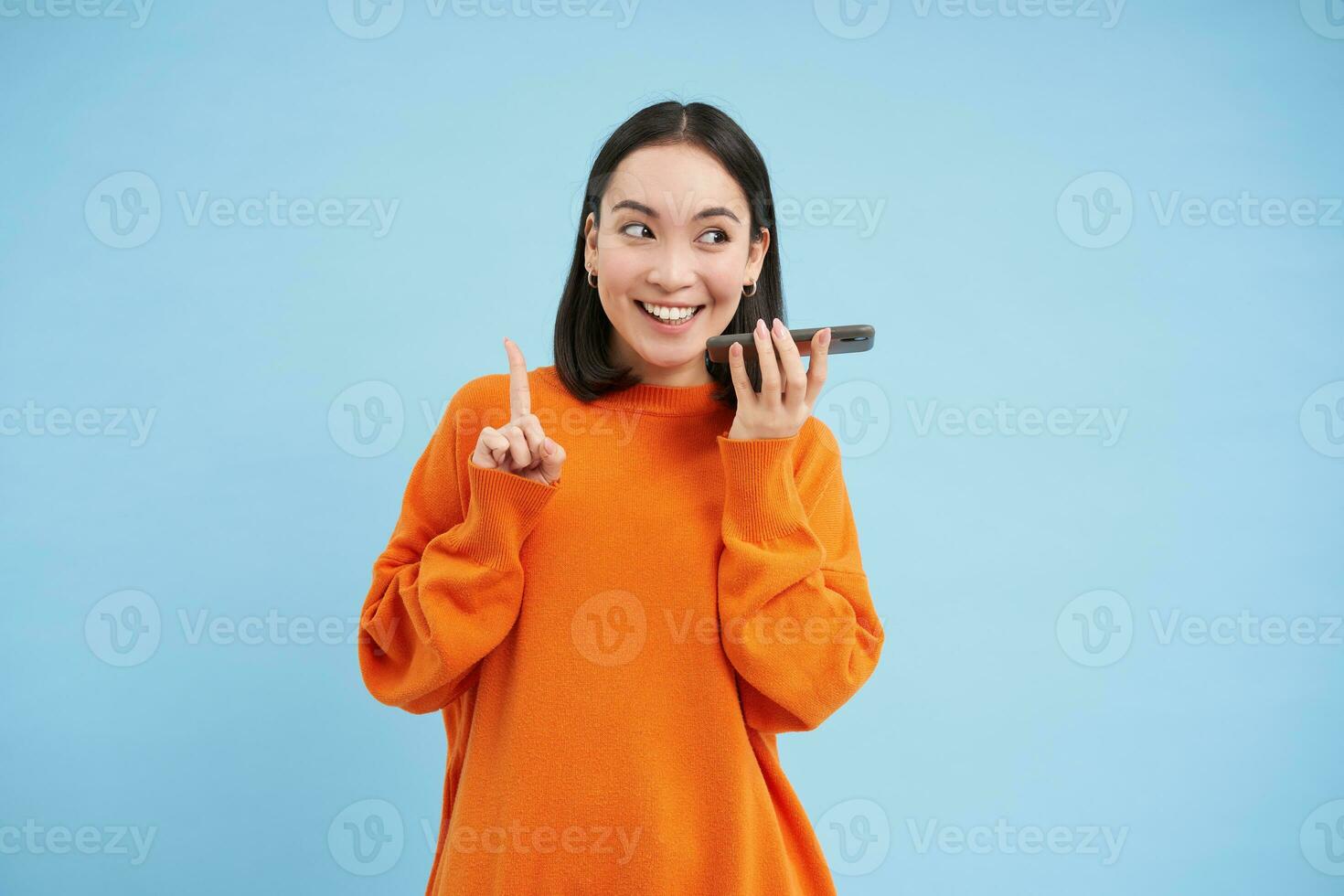 Lycklig asiatisk kvinna uppgifter röst meddelande, upprepningar ord i språk inlärning mobil app, samtal i smartphone högtalartelefon, blå bakgrund foto