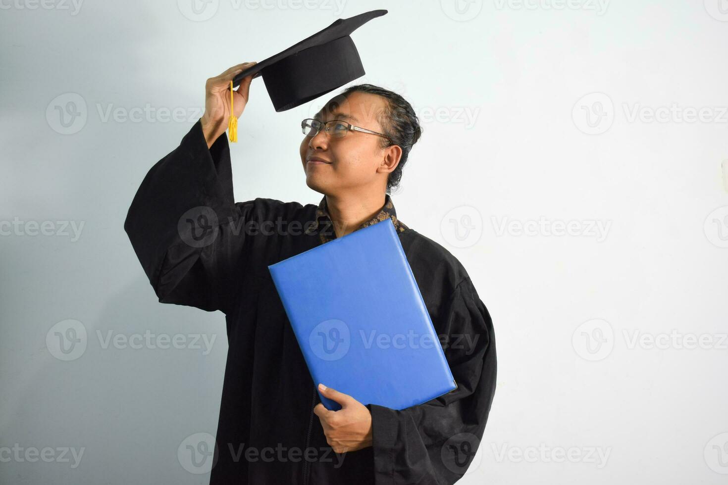 uttrycksfull av vuxen indonesien manlig ha på sig gradering morgonrock, hatt och glasögon, asiatisk manlig gradering föra tom blå certifikat isolerat på vit bakgrund, uttryck av porträtt gradering foto