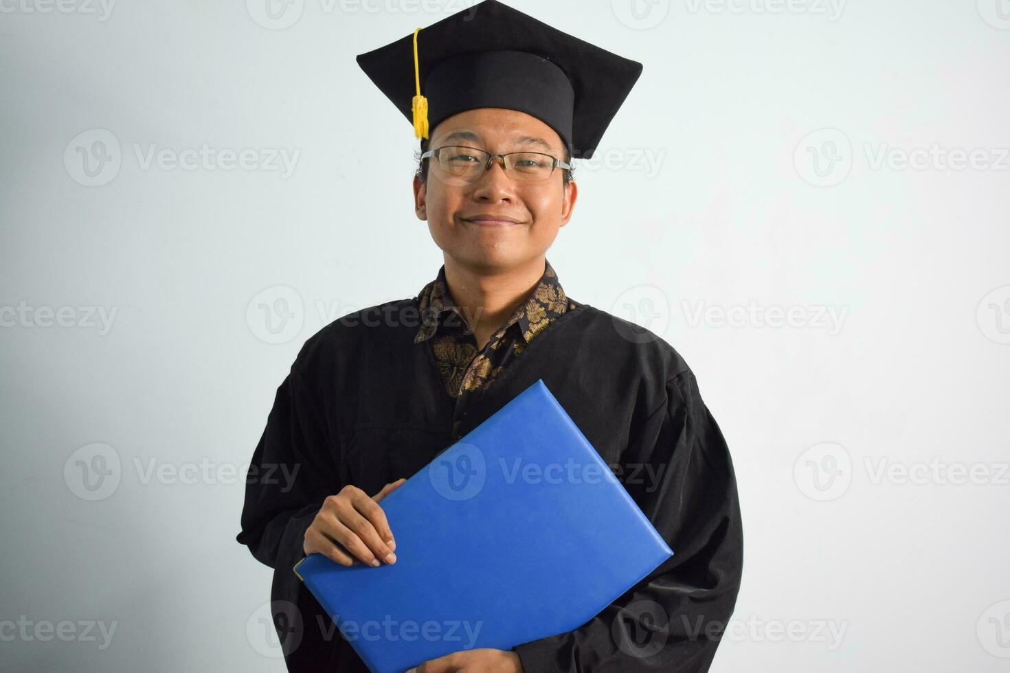 uttrycksfull av vuxen indonesien manlig ha på sig gradering morgonrock, hatt och glasögon, asiatisk manlig gradering föra tom blå certifikat isolerat på vit bakgrund, uttryck av porträtt gradering foto