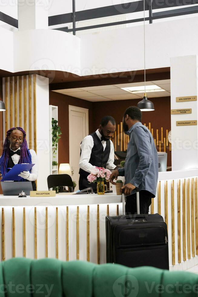 afrikansk amerikan hotell främre skrivbord personal hjälpa gäster på reception, kontroll i turister, begärande för bokning detaljer under checka in. turist med bagage anländer på tillflykt bokning rum foto