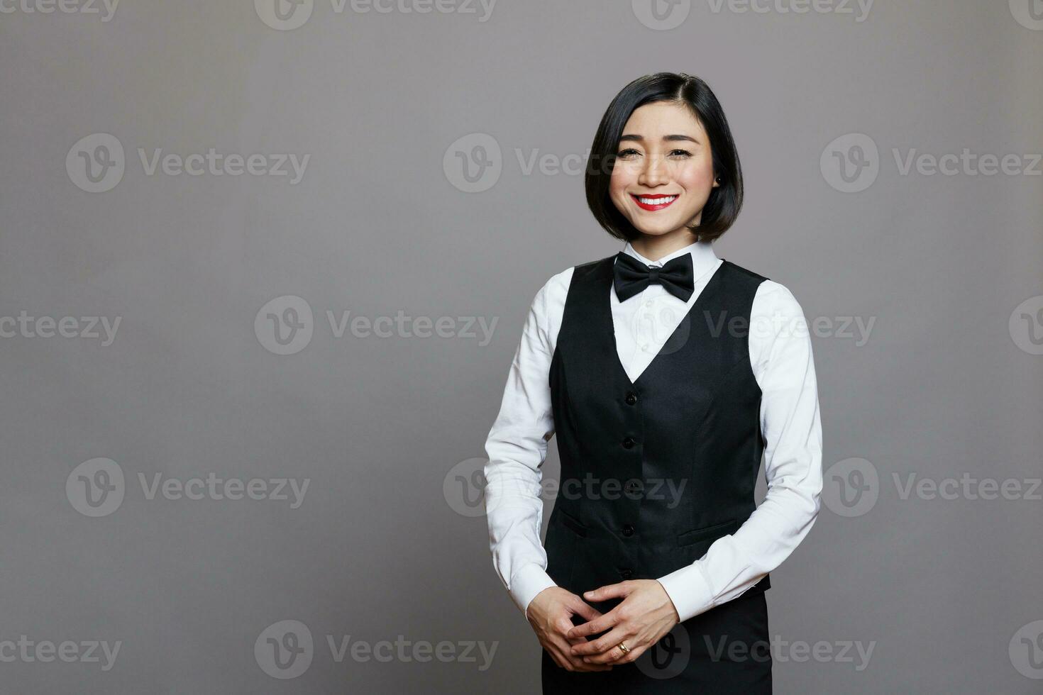 attraktiv leende asiatisk receptionist bär professionell enhetlig stående och ser på kamera med positiv ansiktsbehandling uttryck. ung servitris studio porträtt på grå bakgrund foto