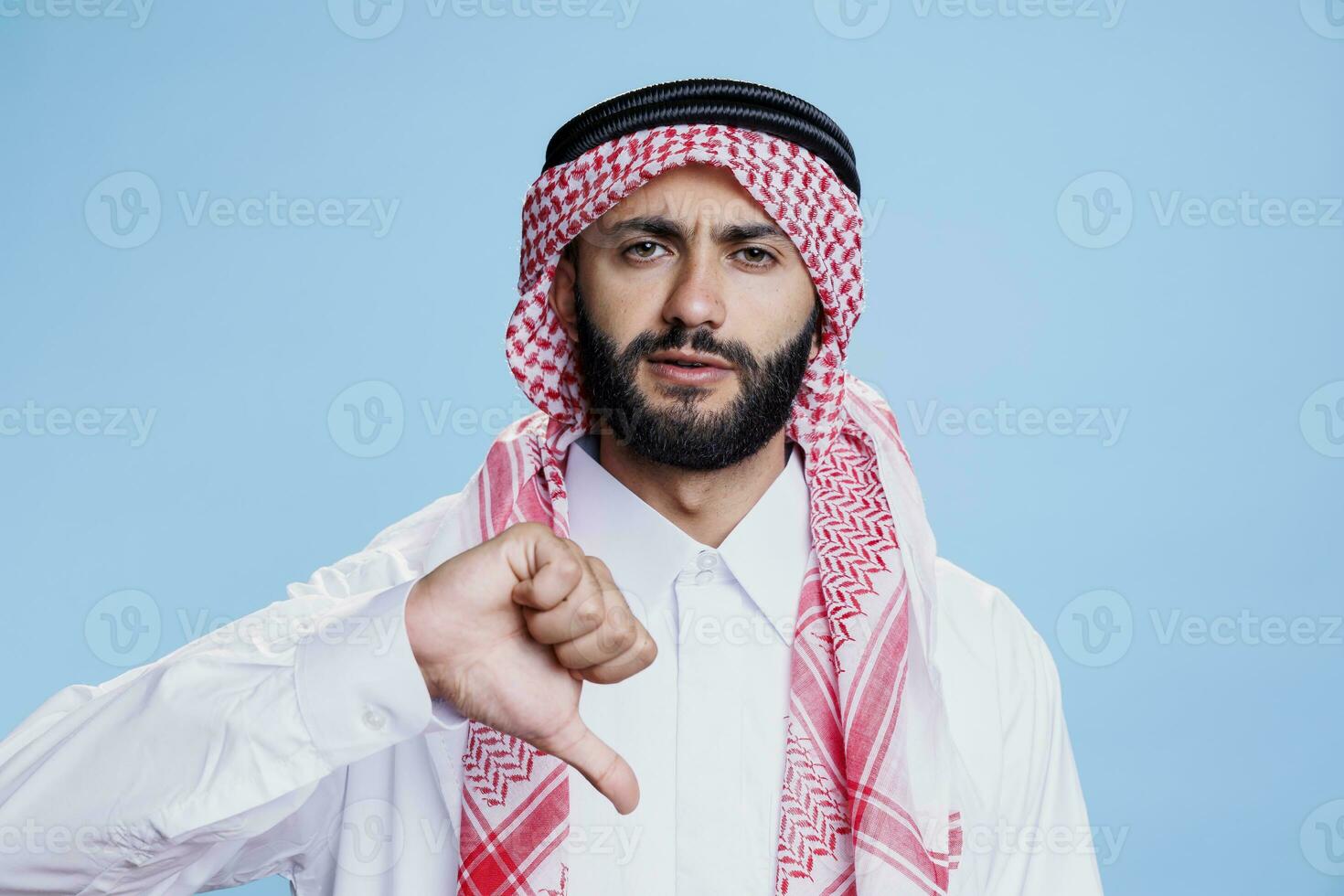 muslim man klädd i traditionell huvudbonad som visar tumme ner negativ gest och ser på kamera. allvarlig arab bär islamic kläder medan Framställ med ogilla tecken porträtt foto