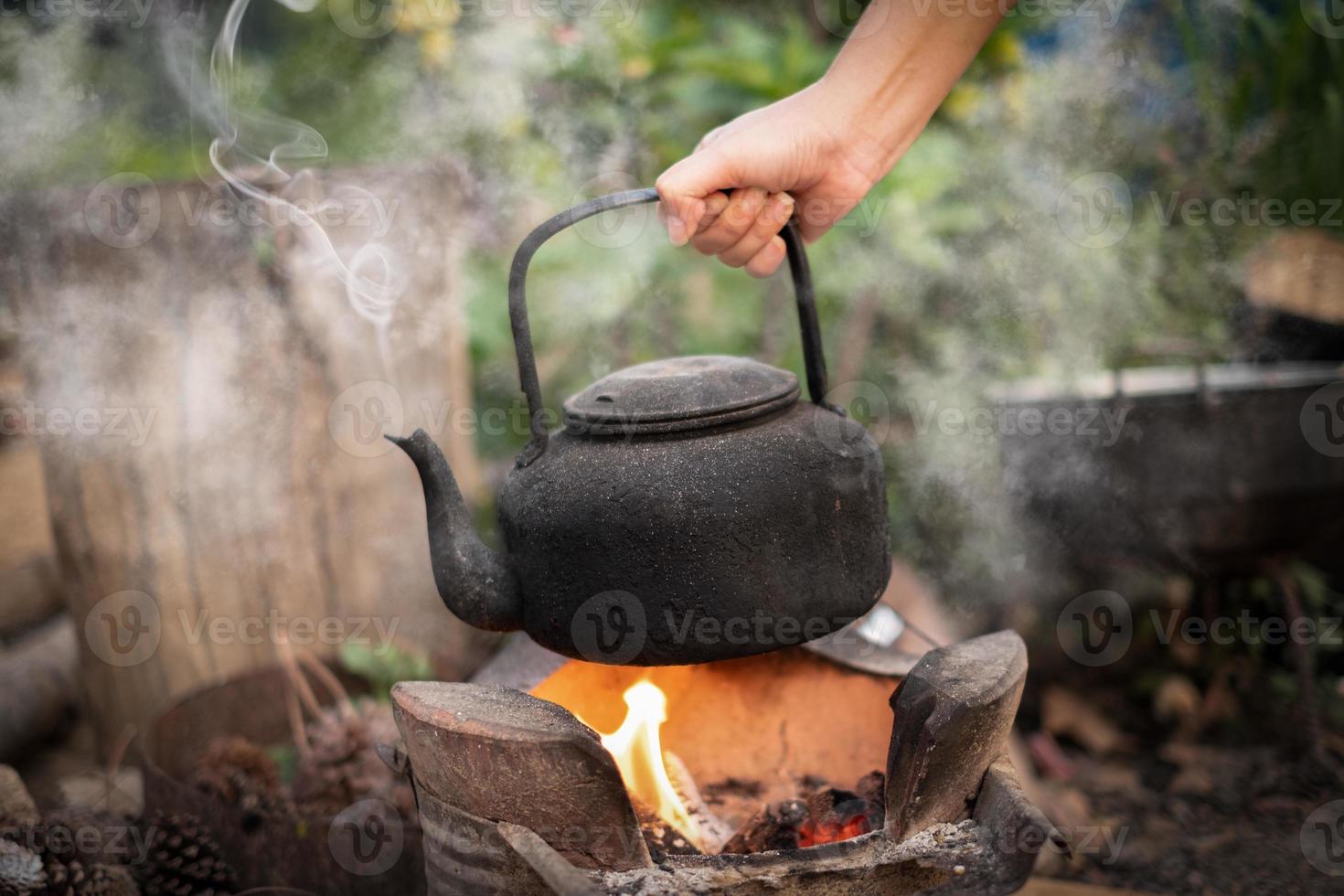 närbild hand hålla koka vatten gammal vattenkokare på elden med en kol spis på suddig bakgrund foto
