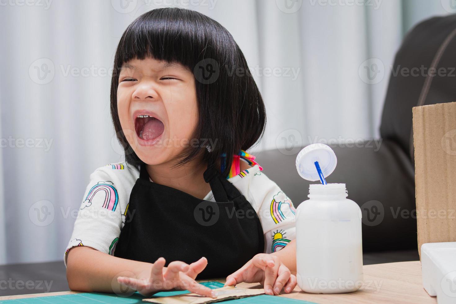 glad asiatisk tjej bär svart förkläde, barn skratt medan han limmar på ett hantverk. begreppet lärande genom lek. barn i åldern 4-5 år. foto