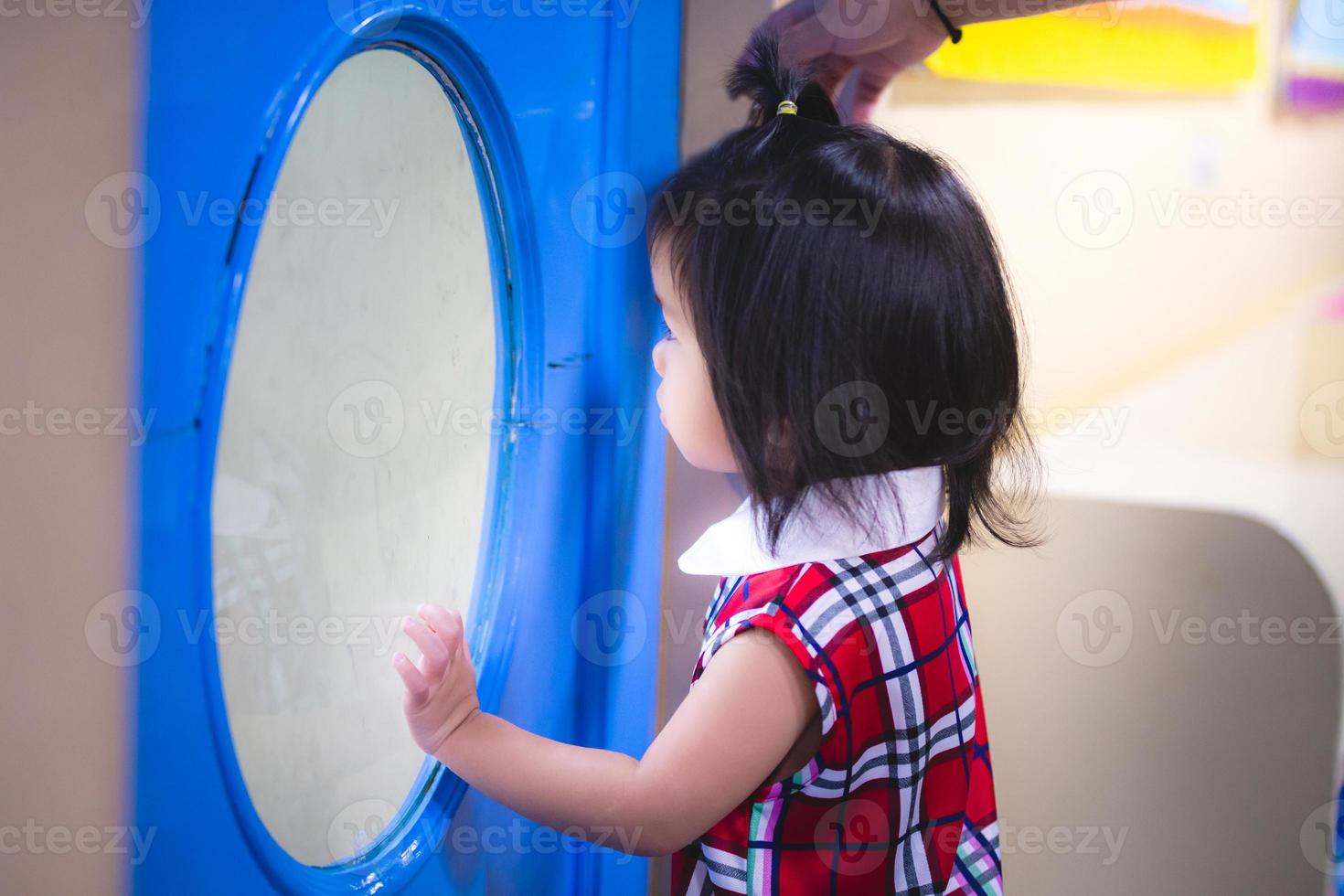 Bangkok, Thailand. 20 juli 2019. asiatiskt barn liten flicka stod genom ett klart glas med ensamt humör och missade. söt tjej klädd i en röd klänning, 2 år gammal. sommarsäsong. foto