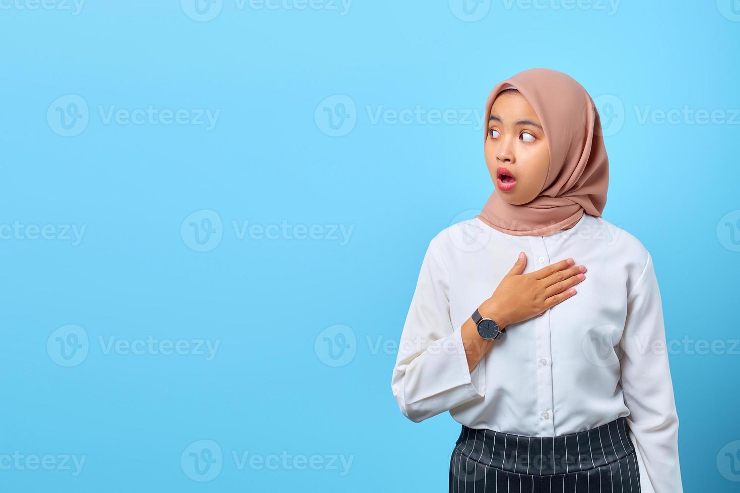 porträtt av chockad ung asiatisk kvinna med öppen mun och lägga handen på bröstet över blå bakgrund foto