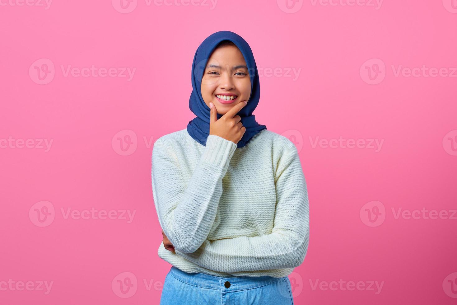 porträtt av leende ung asiatisk kvinna som vidrör hakan med handen på rosa bakgrund foto