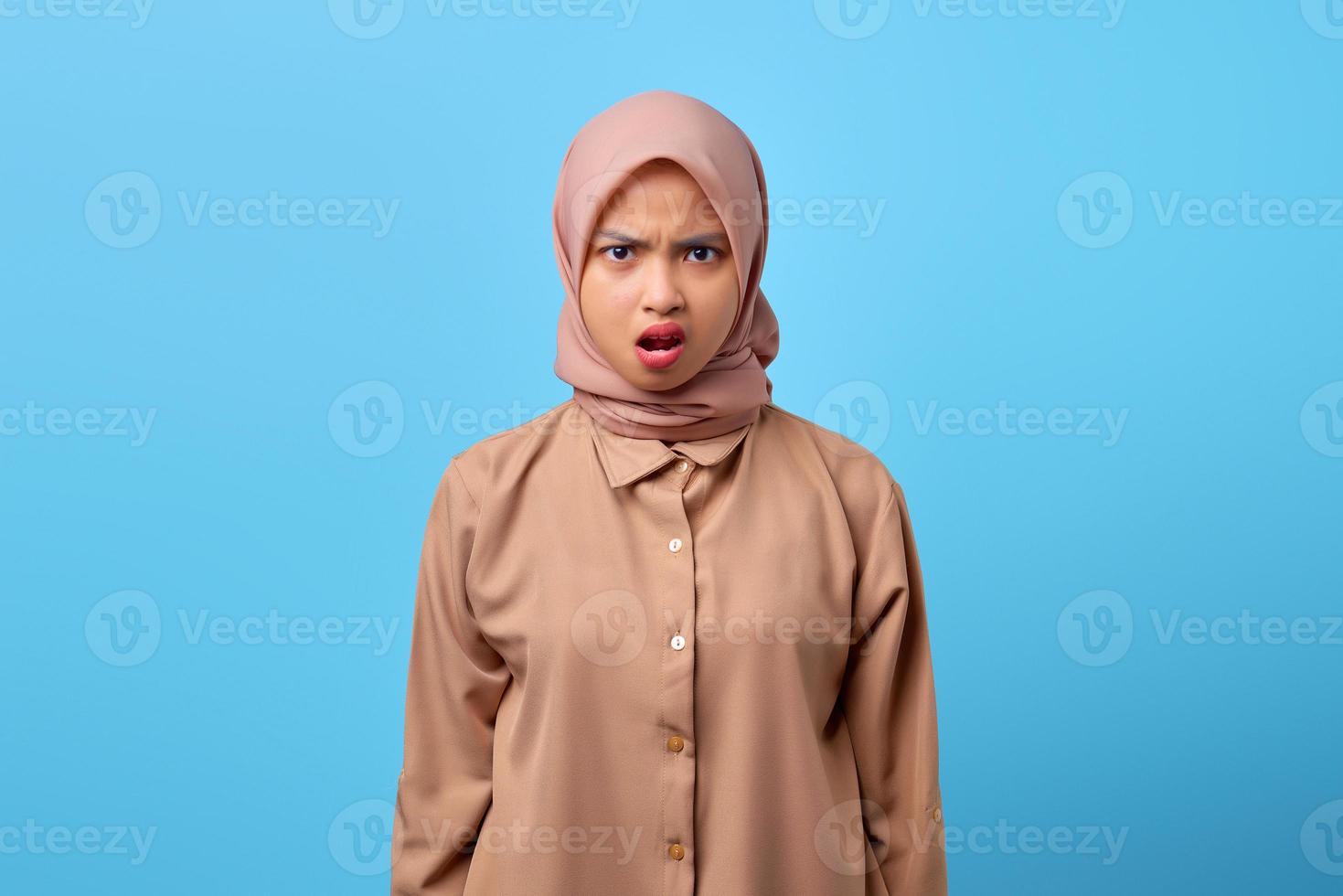 porträtt av chockad ung asiatisk kvinna med öppen mun som bär hijab på blå bakgrund foto