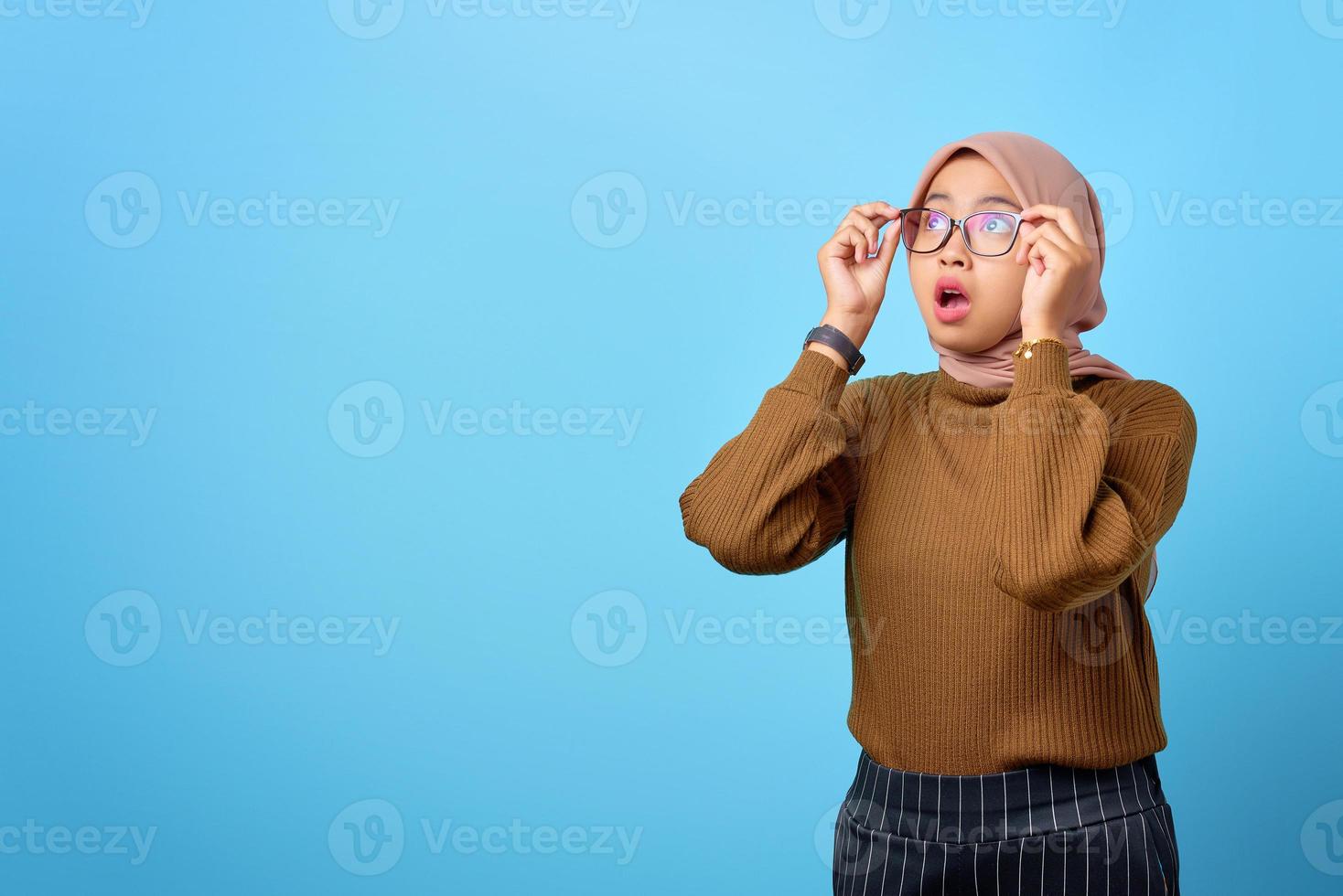 förvånad ung asiatisk kvinna hand på glasögon med öppen mun på blå bakgrund foto