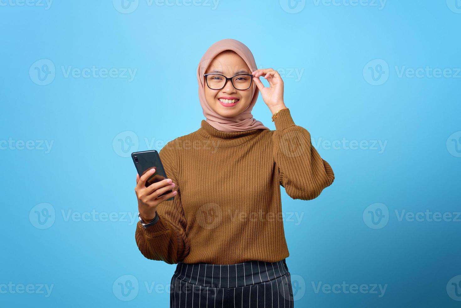 glad ung asiatisk kvinna vidrör glasögon och håller mobiltelefon på blå bakgrund foto
