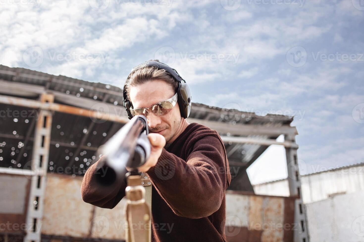 en man i skyddande glasögon och hörlurar. en pumpverkan hagelgevär. däck utomhus. en vägg och en tak med kula hål. sport stänga upp foto