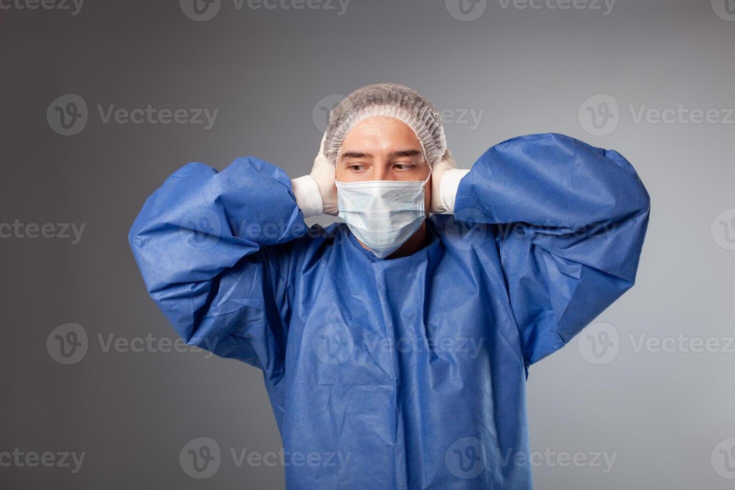 en stilig manlig kirurg är en läkare på en grå bakgrund i de studio. en man i en morgonrock, en medicinsk mask, en hatt. tömma Plats för text. en man omslag hans öron, inte vilja till höra något. foto