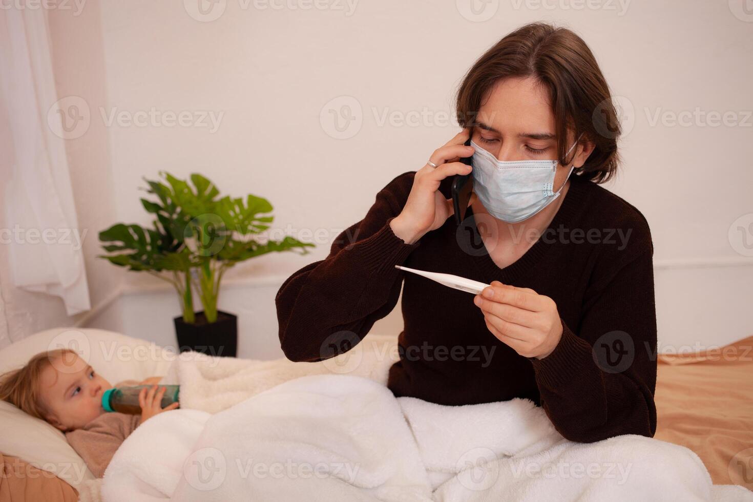 de barn är sjuk, de far i en mask checkar de temperatur av hans dotter. influensa, kall, coronavirus, covid, Hem karantän. de barn är sjuk och lögner på de säng, de man är tar vård foto