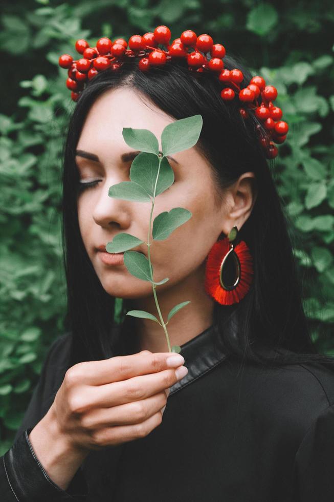 vacker tjej med svart hår och röda tillbehör som håller ett blad i handen nära hennes ansikte mot skogen foto