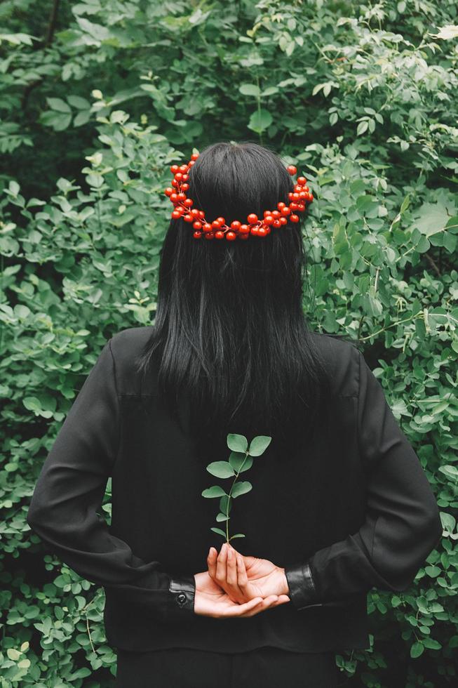 vacker tjej med svart hår och röda tillbehör som håller en kvist i armarna bakom ryggen mot bakgrunden av skogen foto