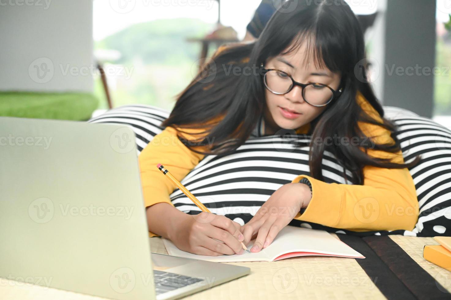 tonårs kvinnlig student sover på kuddar läser bok och använder bärbar dator. hon skriver anteckningar för att förbereda sig för tentamen. foto