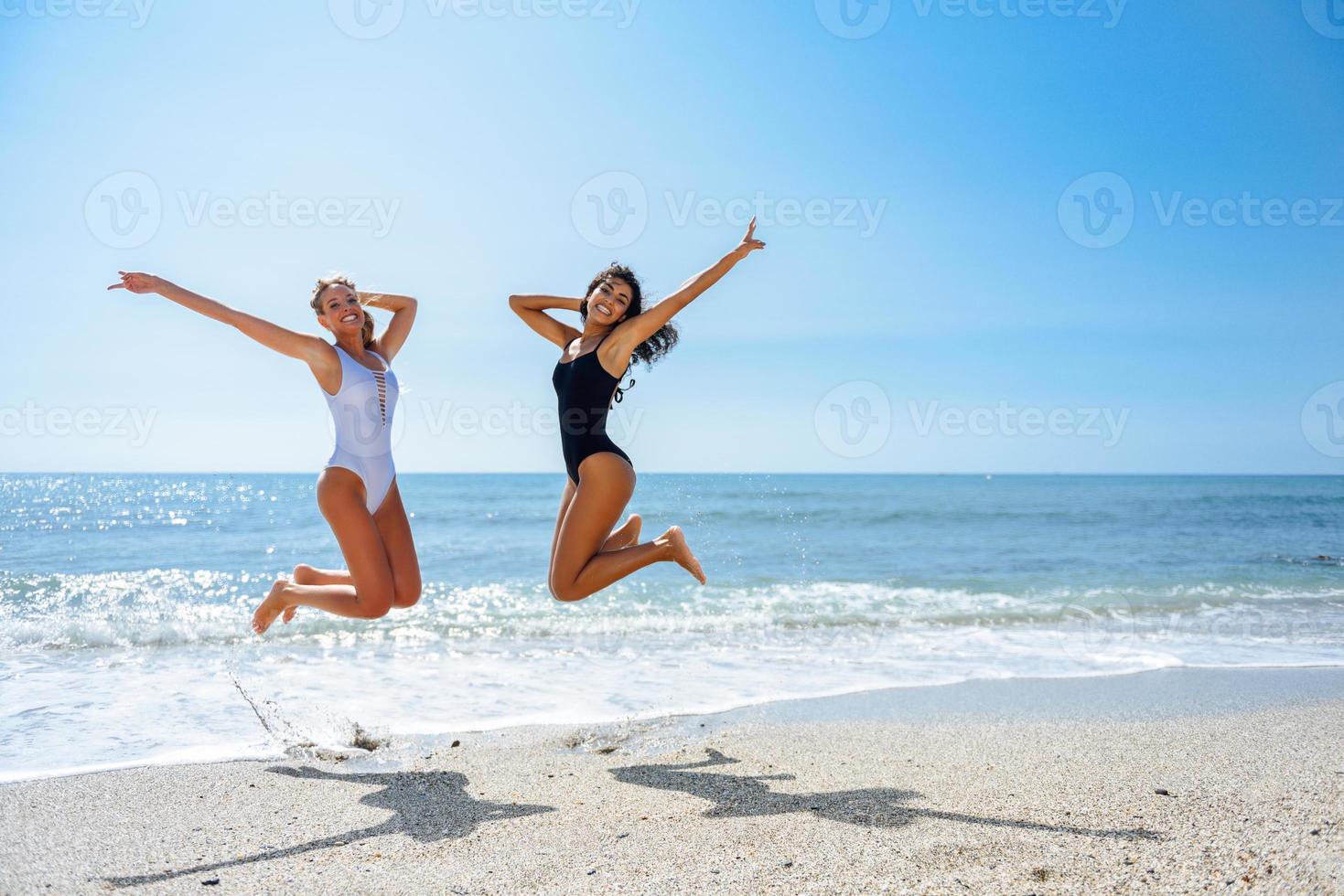 två roliga tjejer i baddräkt som hoppar på en tropisk strand foto