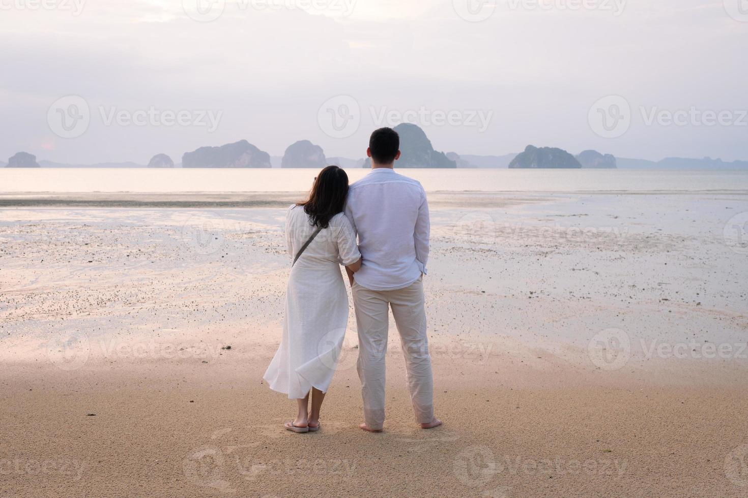 ungt gift par som står på stranden och tittar på solnedgången på smekmånadssemester. samhörighetskoncept foto