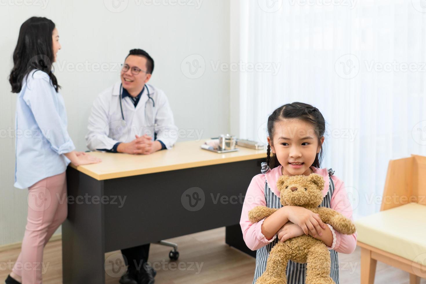 ung asiatisk tjej som håller nalle och står framför mamma och läkare på sjukhusets utvecklingsklinik. hälsovård och medicinsk koncept foto