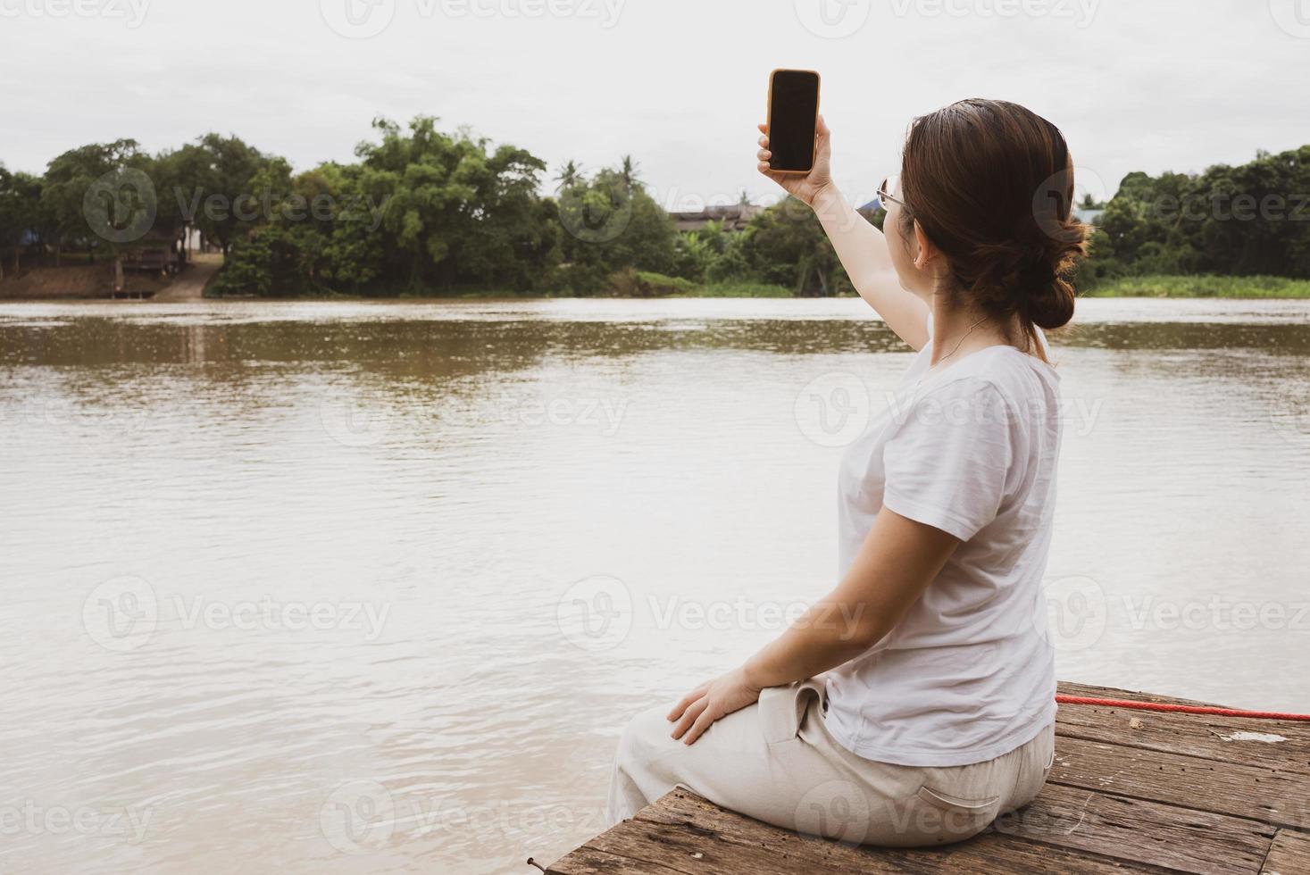 ung asiatisk vuxen kvinna som använder smartphone till selfie under semester vid floden foto