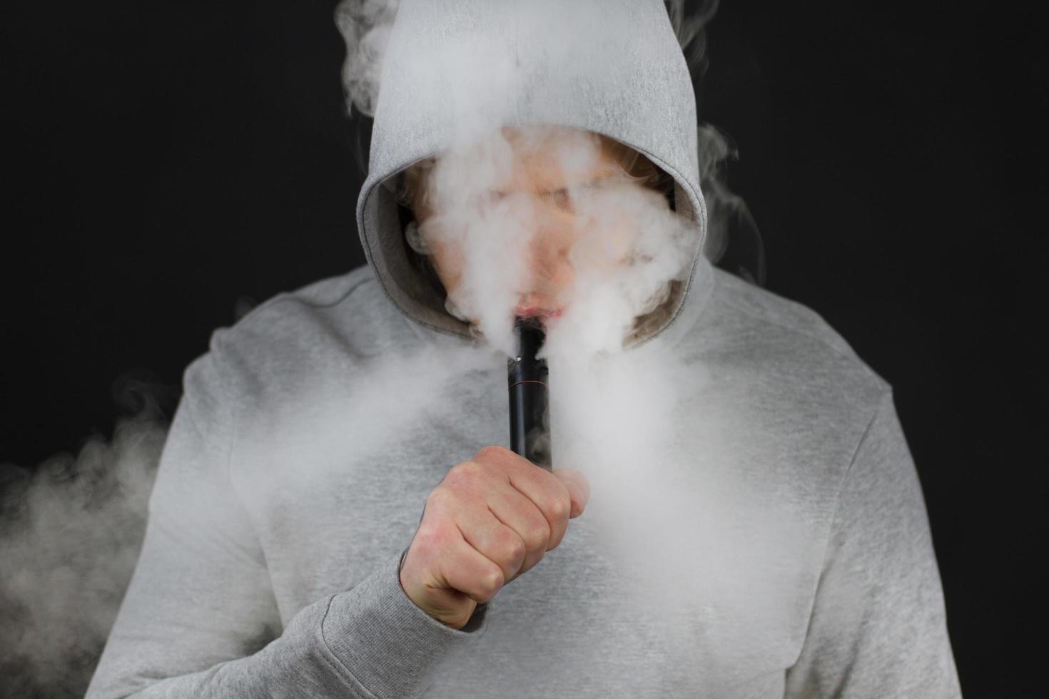 mannen röker en elektronisk cigarett på den mörka bakgrunden, män i huva vaping och släpper ut ett moln av ånga. kille med vaping på svart bakgrund foto