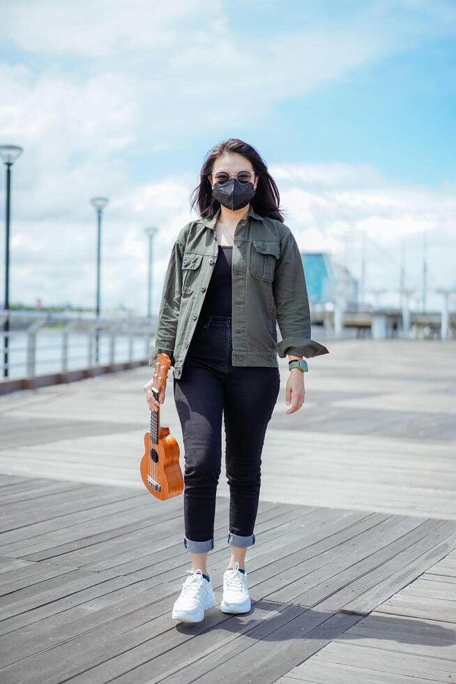 spelar ukulele av ung skön asiatisk kvinna bär jacka och svart jeans Framställ utomhus foto